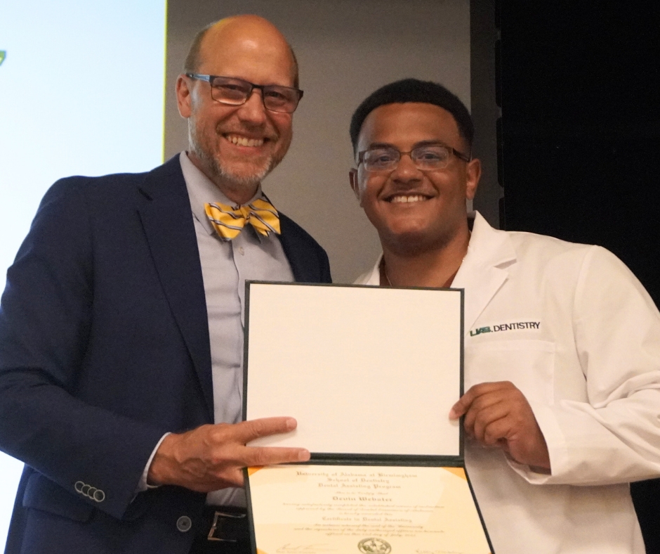 A bald man with glasses in a suit with yellow bowtie standing next to man with black hair and glasses wearing a white coat holding a certificate